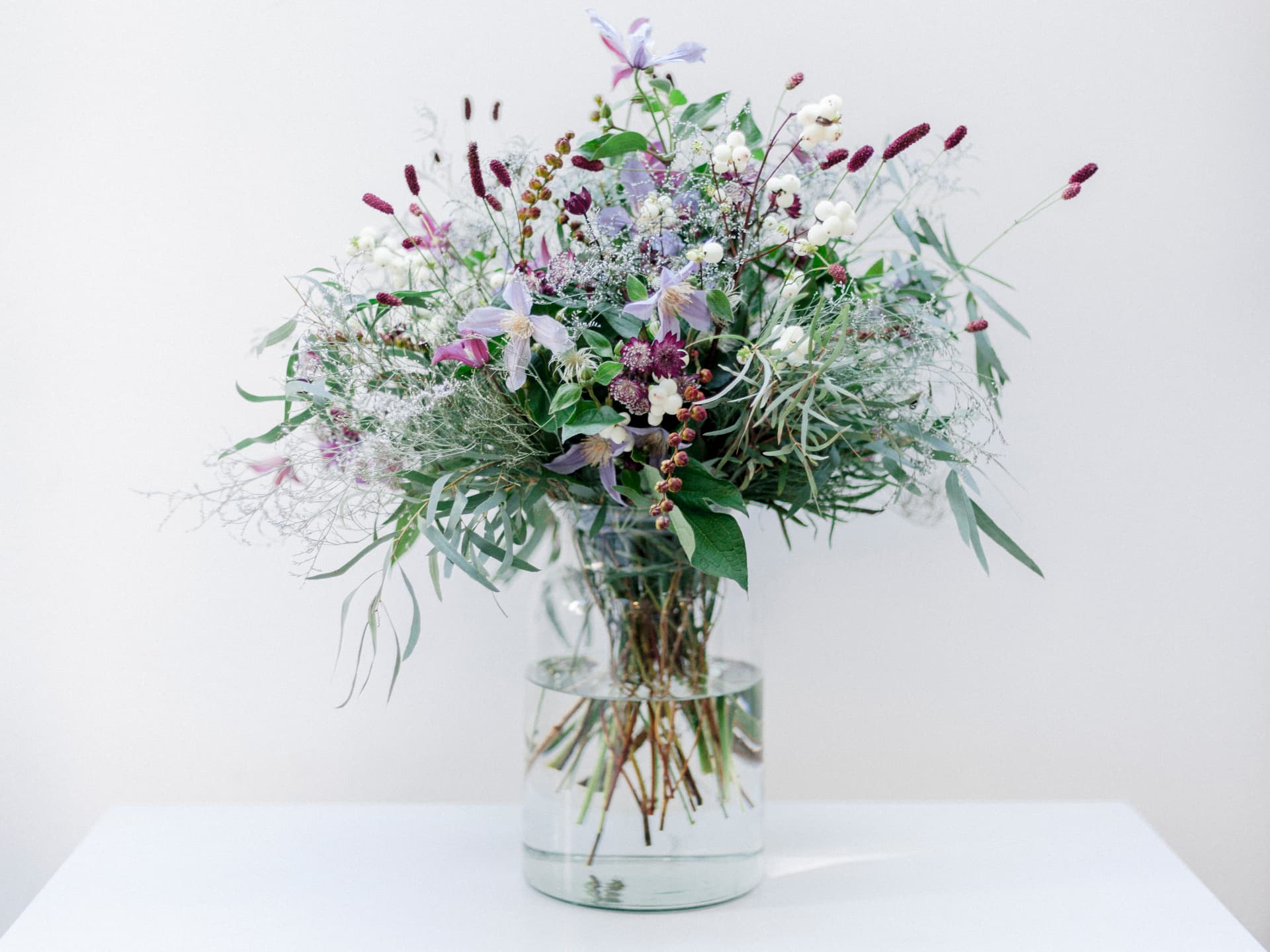 Loose seasonal arrangement with clematis, white berries and burgundy flowers in a glass vase